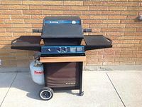 Side and front views of the black Broil King gas barbecue grill showing its functional control knobs, side shelves, and wheels with attached white Flagro propane tank.