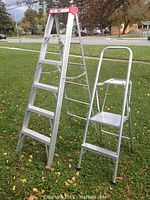 Photo showing both aluminum step ladders standing on grass outdoors. The 6-foot ladder is taller with a red branded top. The shorter platform ladder is next to it with a tubed handle and platform top.