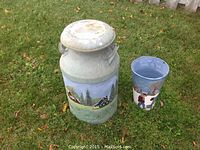 Milk can and sap bucket set displayed outdoors on grass, showing painted rural scenes.