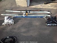 Full view showing two cross country skis with bindings, two pairs of inline skates (white and black), and ski poles lying on concrete.