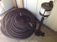 Column sump pump with two long black coiled discharge hoses against a wall corner on a concrete floor.