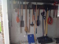 Photo showing multiple garden tools hanging on wall including shovels, rakes, brooms, and the electric trimmer.