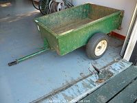 Side view of green steel trailer showing the rectangular cargo bed and hitch arm with some rust and wear