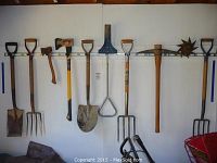 Wide view of all garden tools hanging on white wall rack including shovels, pitchforks, axes, pickaxe, and spiked wheel cultivator.