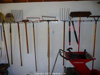 Wide view of assorted garden tools hanging on wall including shovels, rakes, pitchforks, loppers, broom and wheelbarrow on floor.