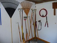 Wide view showing large leaf rakes, hoes, loppers, saw, axe, and other tools hung on wall and leaning against it.
