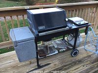 Silver Weber gas grill with black lid, side burner, and attached propane tank on a wheeled cart outside on a wooden deck. Lower storage rack holds grilling accessories.