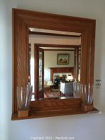 Full view of the beveled edge mirror in a wooden frame with two glass vases attached at the bottom sides. The mirror reflects a room with furniture and a painting.