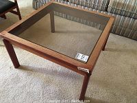 Full view of square coffee table showing wooden frame and glass inset top on carpeted floor with striped sofa background.
