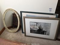 Lot with oval framed mirror and two framed art prints leaning against wall on carpeted floor. Mirror frame is ornate cream-colored. Prints have wood and blue-green frames.