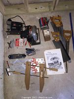 Overview photo showing collection of woodworking tools including jigsaw, circular saw, hand saws, clamps, woodworking books, measuring tools and accessories arranged on a concrete floor.
