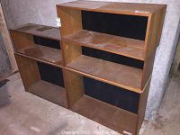 Photo showing all 5 modular wooden shelves grouped together on a floor in a dimly lit basement or storage area, all with a natural wood finish and black back panels, showing surface dust and wear.