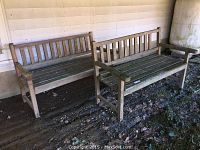 Two matching outdoor wooden benches positioned side by side under a covered area. Both tables show heavy weathering and moss growth.