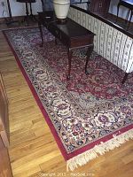 Full length view of the rug under furniture showing rich red border and detailed floral and vine patterns.