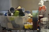 Plastic tub on table filled with various plastic containers, red solo cups stacked in background, and multiple plastic glasses and cups visible in foreground