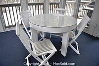Photo showing the round wooden table with glass top and the four folding wooden chairs arranged around it on a porch area.