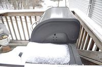 Side view of black BBQ grill covered partly in snow showing curved lid and left metal shelf with chipping.