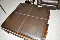 Photo showing top view of square ottoman coffee table with dark brown leather cushion divided into four sewn sections and wooden frame base.