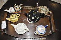Six decorative teapots arranged on a metal serving tray with wooden handles showing varied designs and color schemes.