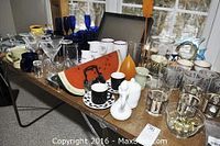 Wide view of assorted glasses and decor items on table including milk glass, blue glasses, watermelon tray, and other decorative glasses.