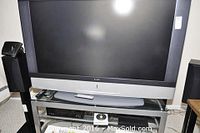 Sony 60-inch LCD projection television on glass and metal stand with two speakers. View shows front of the TV with a silver bottom bezel and Sony logo, and media devices below on glass shelves.