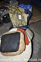 Several dog beds stacked including a beige plush bed with black center, round cream bed with orange accents, and tan quilted bed, with blue plastic double dog bowl behind and stacked plastic bins.