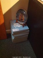 Stack of seven blue boxes labeled 'Legendary Ships of the Seas' with one displayed plate on wooden stand showing a painted sailing ship at sea with clouds and sunset colors.