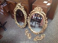 Two ornate gold-painted wall mirrors leaning against furniture with matching pair of gold wall sconces placed in front, showing design details and sizes