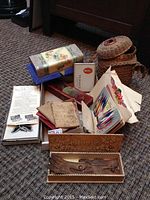 Overall view showing large woven basket, floral metal tin, red floral folding box, carved glove box, hymnals, and assorted paper items on a carpet.