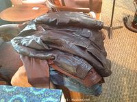 Four men's jackets stacked piled on a chair and furniture, showing leather and darker materials.