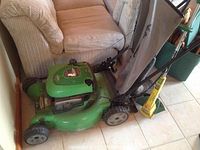 Green LawnBoy gas-powered lawnmower with attached grass collection bag, placed indoors by a couch.