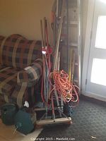 Photo showing assorted garden tools including shovels, brooms, extension cord, watering can, bird feeder, and a couch in the background.