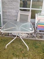 Square glass-top patio table 36x36 with metal frame, placed on grass near stones and wall.