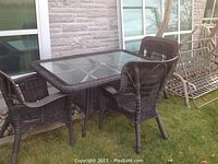 Patio set with rectangular wicker-framed glass table and three wicker chairs on lawn near house exterior.
