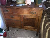 Front view of oak serpentine dresser showing three drawers on the left and one cabinet door on the right with brass handles. Surface shows wear and some staining.