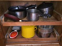 Full view of shelf inside cabinet holding various pots and pans including copper bottom pan and yellow enameled pot.