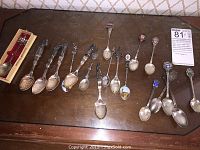 Collection of 16 souvenir spoons arranged on a wooden surface, showing different designs and origins.