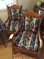 Two wooden armchairs with floral seat and back cushions, placed next to each other inside a room showing overall wear.