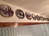 Wall-mounted collection of ten Western collector plates by artist Chuck Ren showing horses, cowboys, and Native Americans.