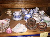 Overview of assorted china including tea cups, saucers, plates, and gravy boat arranged on a wooden shelf.