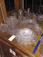 View of multiple cut glass serving pieces including bowls, decanters, pitchers, lidded candy dishes on a wooden surface in front of a window.