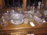 Full lot of cut glass serving pieces arranged on a wooden surface, including decanters, bowls, candlestick, covered dishes, and plates.