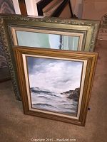 Two framed paintings stacked; one with a gold frame showing ocean waves and rocky shoreline, the other partial framed art behind it