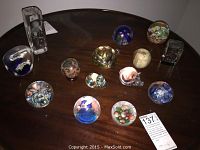 Top-down view of 13 assorted glass paperweights and vase on wooden table.