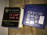 Two hardcover stamp collector albums on floor. One titled The Master Global Stamp Album, another Lotta Deluxe Worldwide Stamp Album in blue.