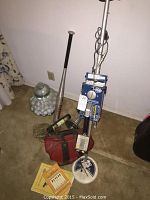 Lot photo showing White's metal detector, aluminum baseball bat, glass jar with golf balls, bowling ball, metal weight, and red bag on floor.