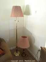 Full view of both gold finished floor lamps with pleated fabric shades, one tall and one shorter, situated near a couch in a room corner.