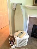 Ironing board leaning against the wall next to fireplace with laundry hamper and bathroom scale on top