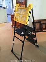 Full image of the black metal step ladder with yellow plastic top tray, showing paint splatters and wear.
