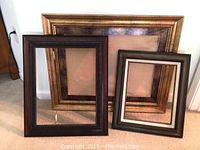 Three wooden picture frames, largest gold tone frame at back, two smaller dark wood frames placed inside, visible decorative molding, standing on carpet against wall.